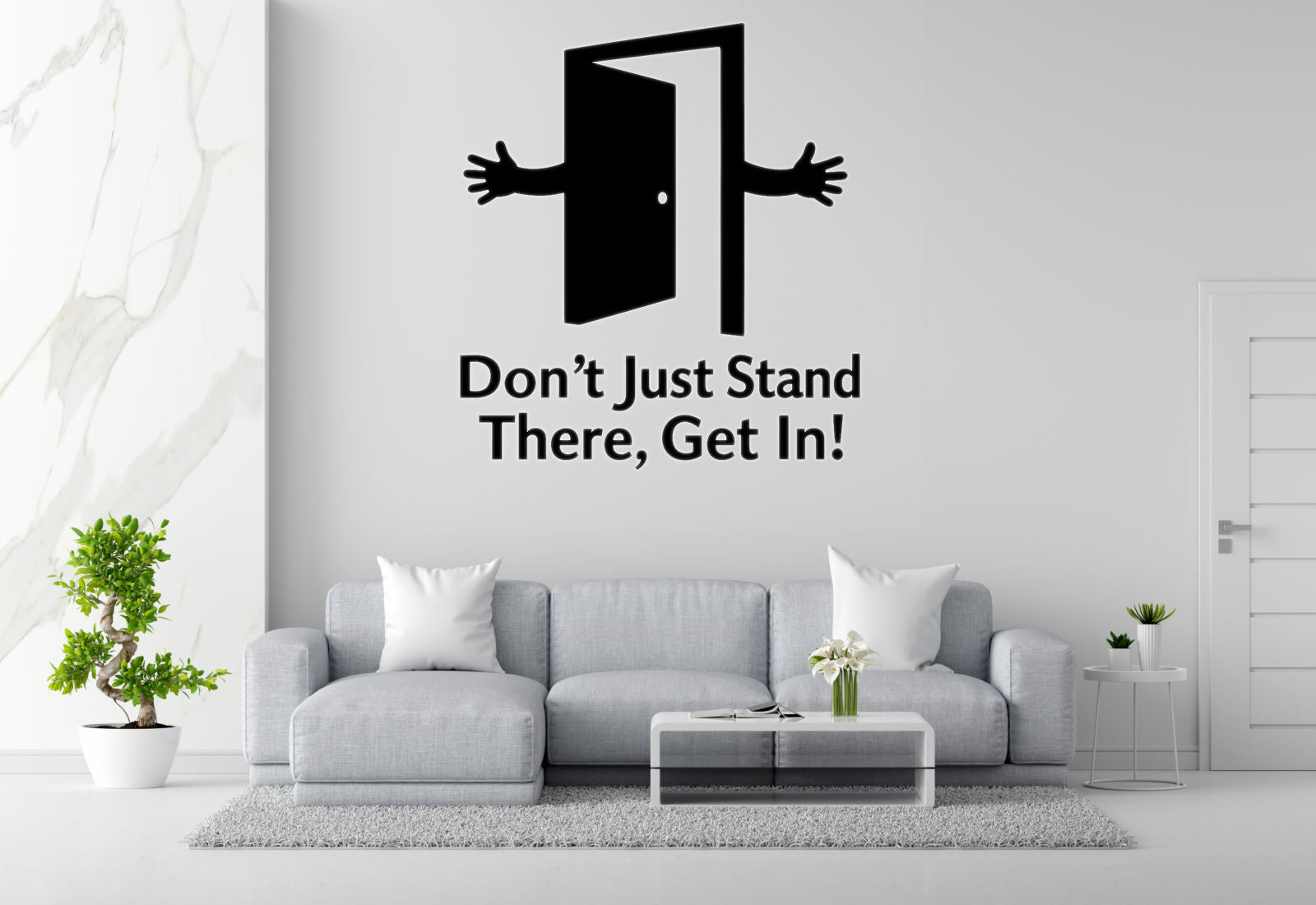 a living room with a gray sofa, a coffee table, and a potted plant. Above the sofa, there is a wall decal with an open door and a hand reaching out, accompanied by the text "Don't Just Stand There, Get In!"