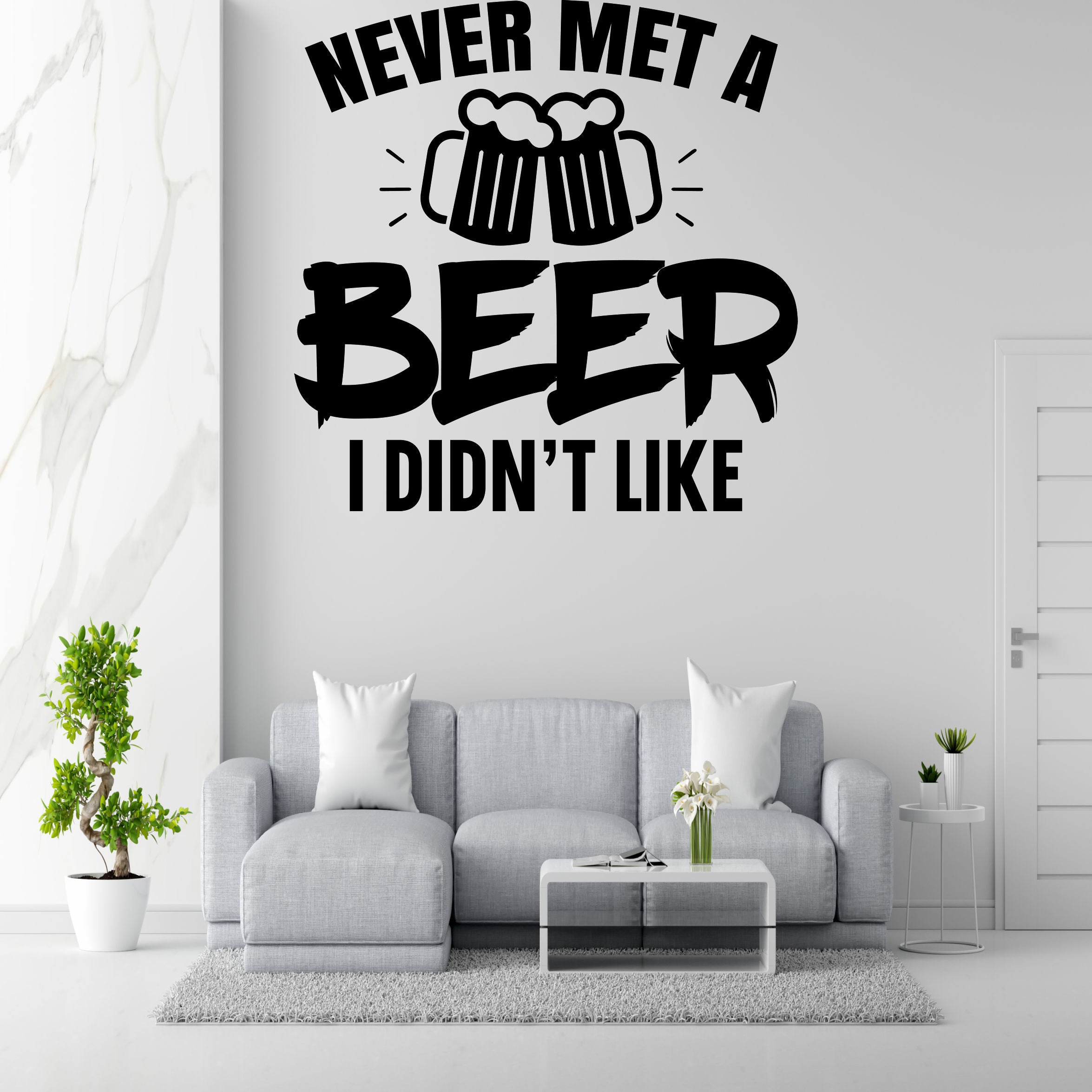 a wall with a humorous text that reads "NEVER MET A BEER I DIDN'T LIKE" in bold letters. Below the text, there are two beer mugs with a celebratory design. The wall is set against a white marble background, and there is a gray couch with two white pillows in the foreground. A coffee table is also present in the room.