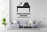 a living room with a gray sofa, a coffee table, and a potted plant. Above the sofa, there is a wall decal that says "WHERE THE MAGIC HAPPENS (AND THE LAUNDRY PILS UP)" in bold letters.