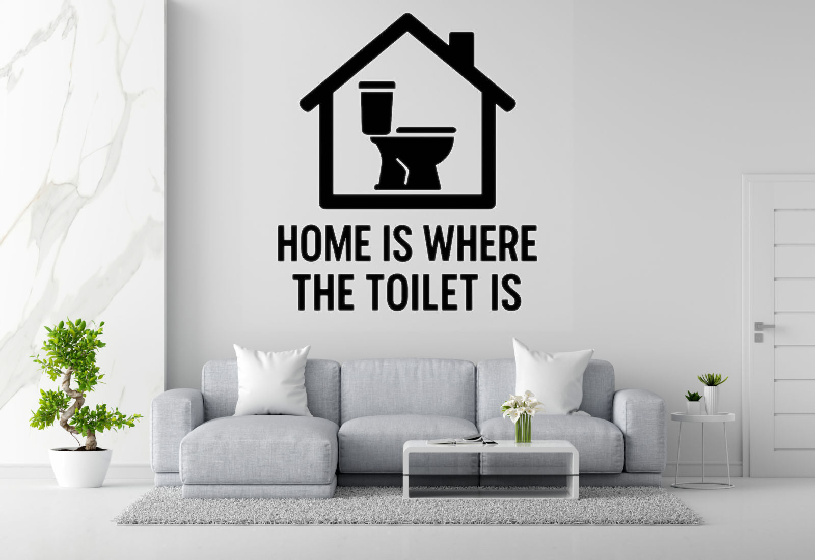 a living room with a gray sofa, a coffee table, and a potted plant. Above the sofa, there is a wall decal that says "HOME IS WHERE THE TOILET IS".