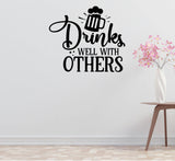 a wall with a black and white text sticker that says "Drinks well with others" above a beer mug icon. Next to the sticker, there is a small white vase with pink flowers.