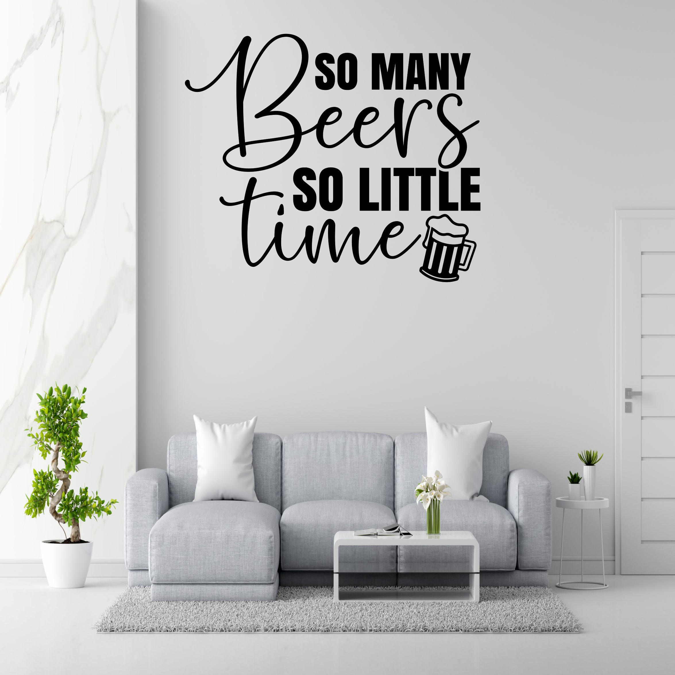 a living room with a gray sofa, a coffee table, and a potted plant. Above the sofa, there is a wall decoration that says "So many beers so little time".