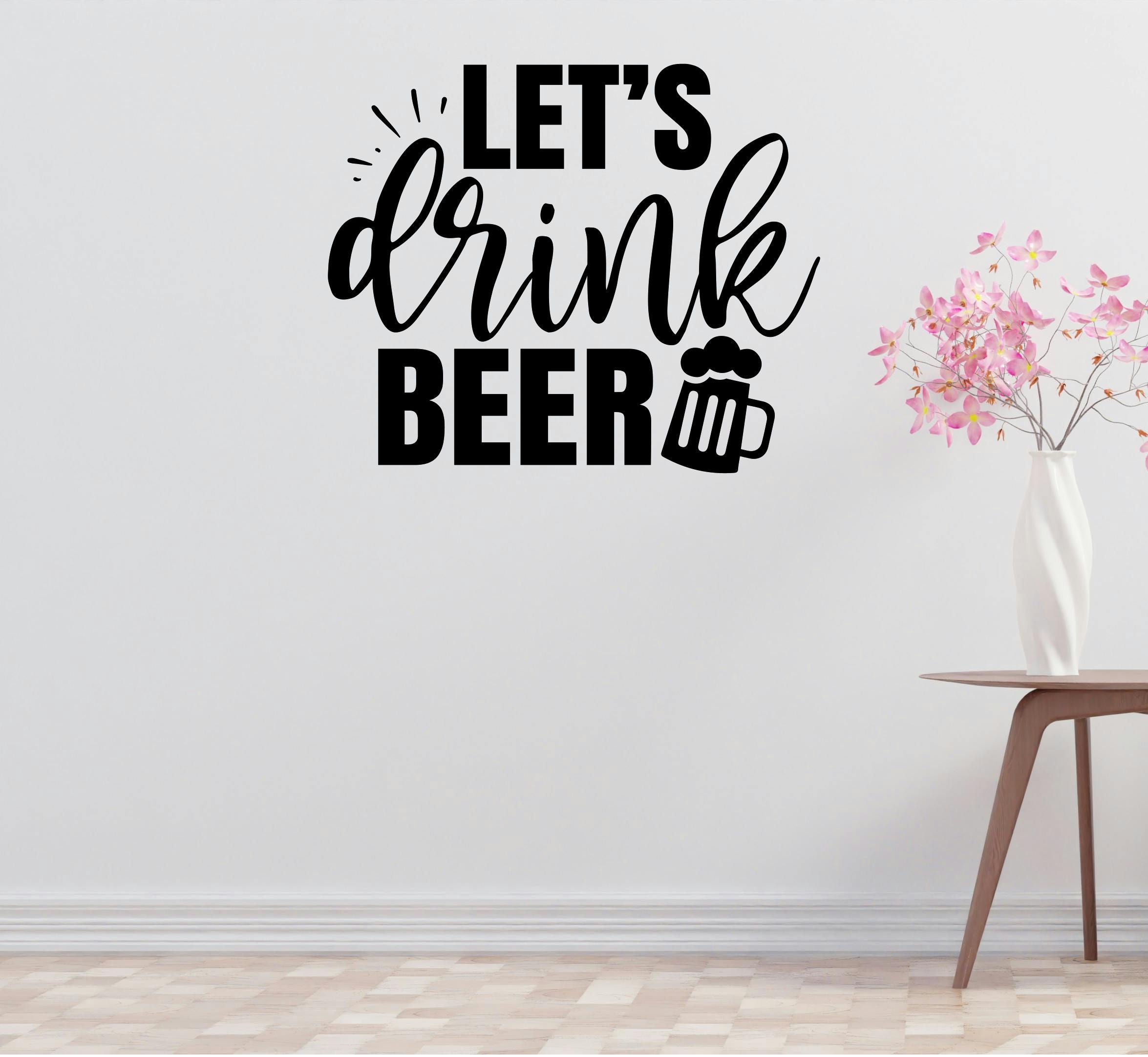 a wall with a black and white text design that says "LET'S DRINK BEER" along with a beer mug icon. Next to the wall, there is a wooden table with a vase of pink flowers on it.