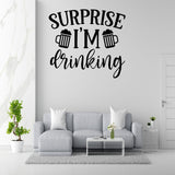 a living room with a gray sofa, a coffee table, and a potted plant. Above the sofa, there is a wall decal that says "Surprise I'm drinking".