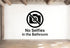 a sign that reads "No Selfies in the Bathroom" with a symbol of a camera crossed out, indicating that photography or selfie-taking is not allowed in that area.