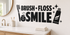 BRUSH FLOSS SMILE black vinyl bathroom wall decal with toothbrush, floss, toothpaste, and sparkles above a pale oak vanity.