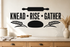 KNEAD RISE GATHER black bakery wall decal with rolling pin, wheat, and bread loaf above a warm baking counter.