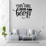 a living room with a gray sofa, a coffee table, and a potted plant. Above the sofa, there is a wall decal that says "This girl needs a beer".